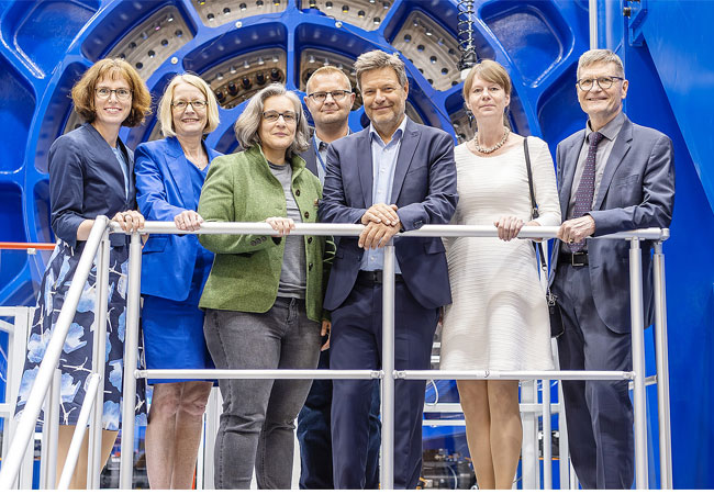 PB: Gruppenbild vor großer Maschine (v.l.n.r.): Dr. Daniela Brönstrup (BMWK), Prof. Dr. Cornelia Denz (Präsidentin der PTB), Dr.-Ing. Julia Hornig (Geschäftsführerin des Kompetenzzentrums Windenergie und Leiterin des PTB-Fachbereichs „Gase“), Holger Kahmann (Konstrukteur und "Erdenker" der Maschine), Bundeswirtschaftsminister*in Dr. Robert Habeck, Dr. Annette Röttger (Mitglied des Präsidiums), Dr.-Ing. Frank Härtig (Vizepräsident der PTB). 