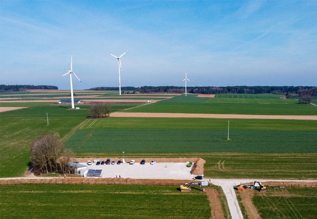 PB: Bei der Baustelleneinrichtung wurde auf dem benachbarten Grundstück der Mutterboden abgetragen und zwischengelagert. Anschließend wurde die Fläche mit einem Schutzvlies aus-gelegt, durch das der Schotter nach Bauschluss sauber entfernbar ist. Im Hintergrund (v. l. n. r.) zu sehen sind drei ältere Windenergieanlagen der Hersteller NEG Micon, Vestas und eine Enercon der Firmengruppe WINDPOWER. / ©: WINDPOWER